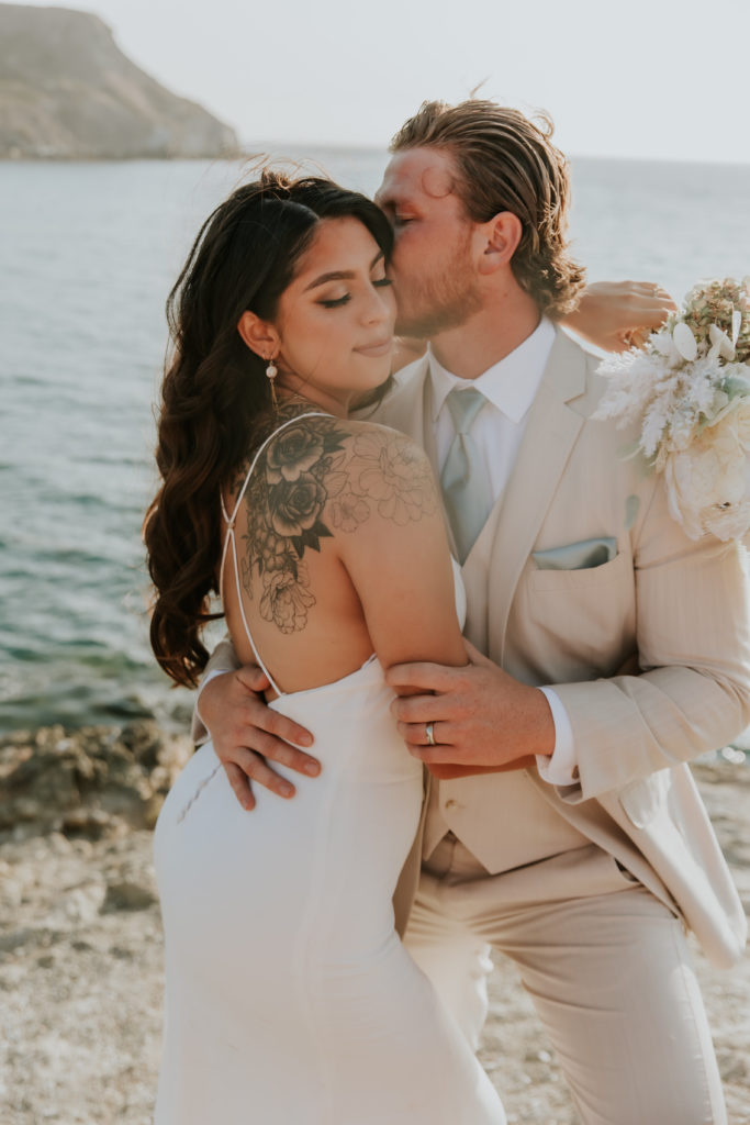 Groom kissing bride on the cheek with the view of Catalina Island Beach in the background