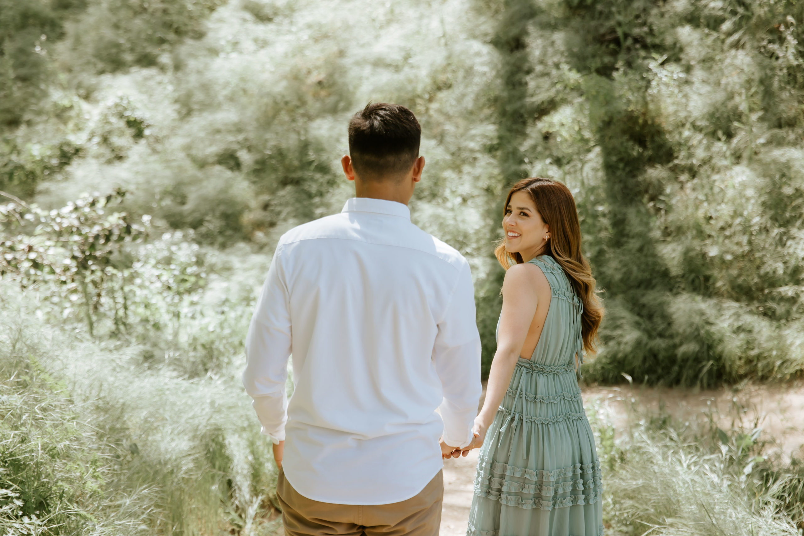 couple walking down a path through the forest during their engagement session and fiancee looks back at her partner