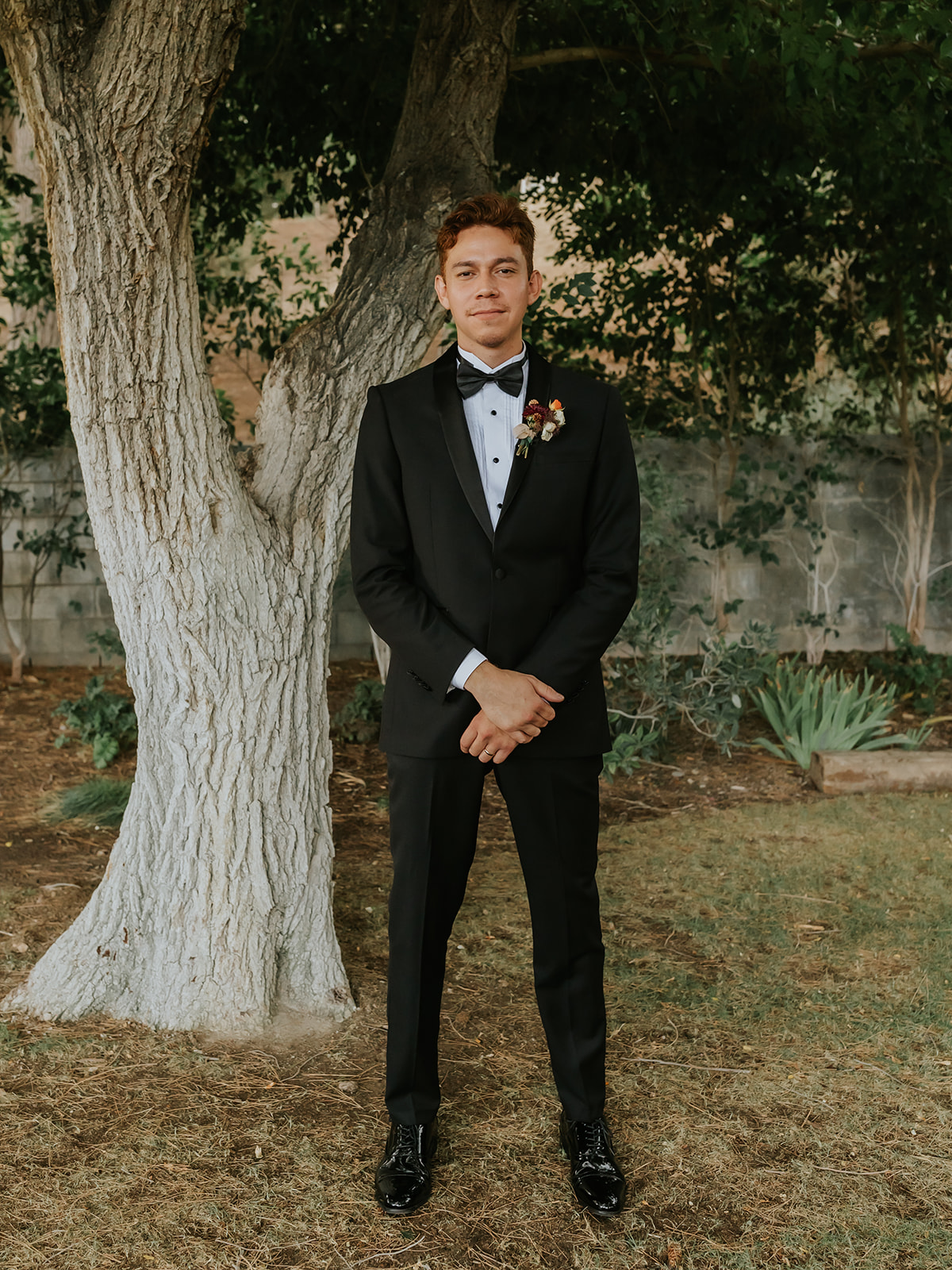 groom standing facing the camera under a tree in his tux smiling
