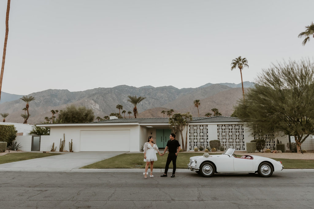 couple holding hands with a classic car and vintage house in the background