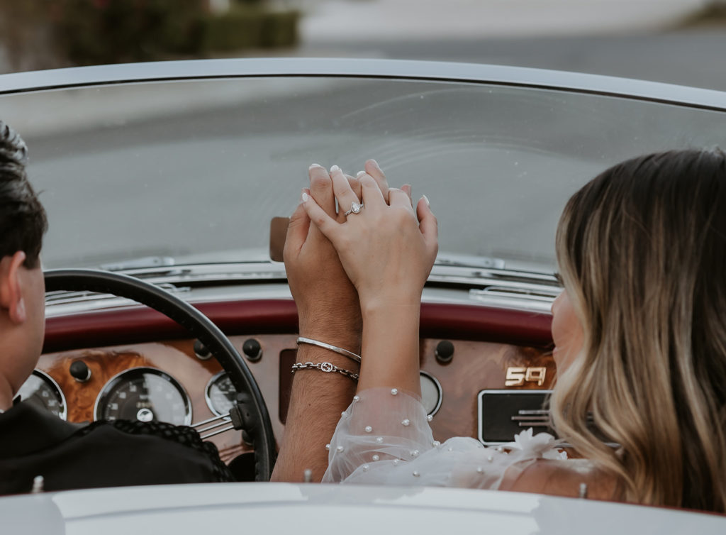 couple holding hands showing off the girls new engagement ring while sitting in a classic car
